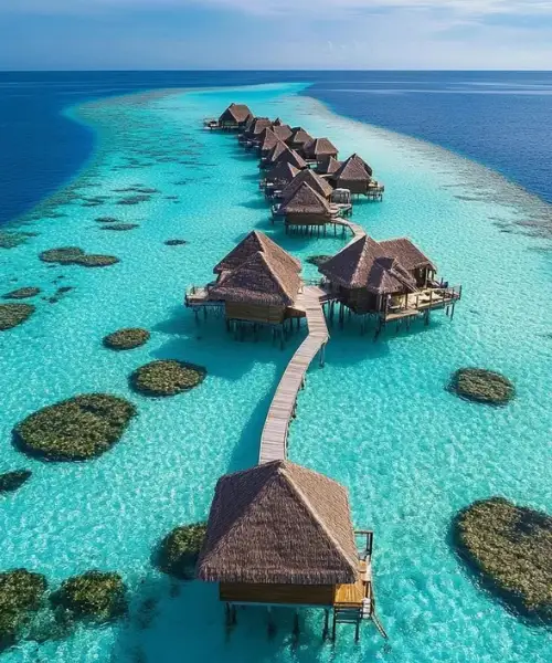 Aerial view of Maldives Water Villa over crystal-clear turquoise lagoon, featuring a row of overwater bungalows connected by a wooden walkway above vibrant coral reefs in the Indian Ocean.